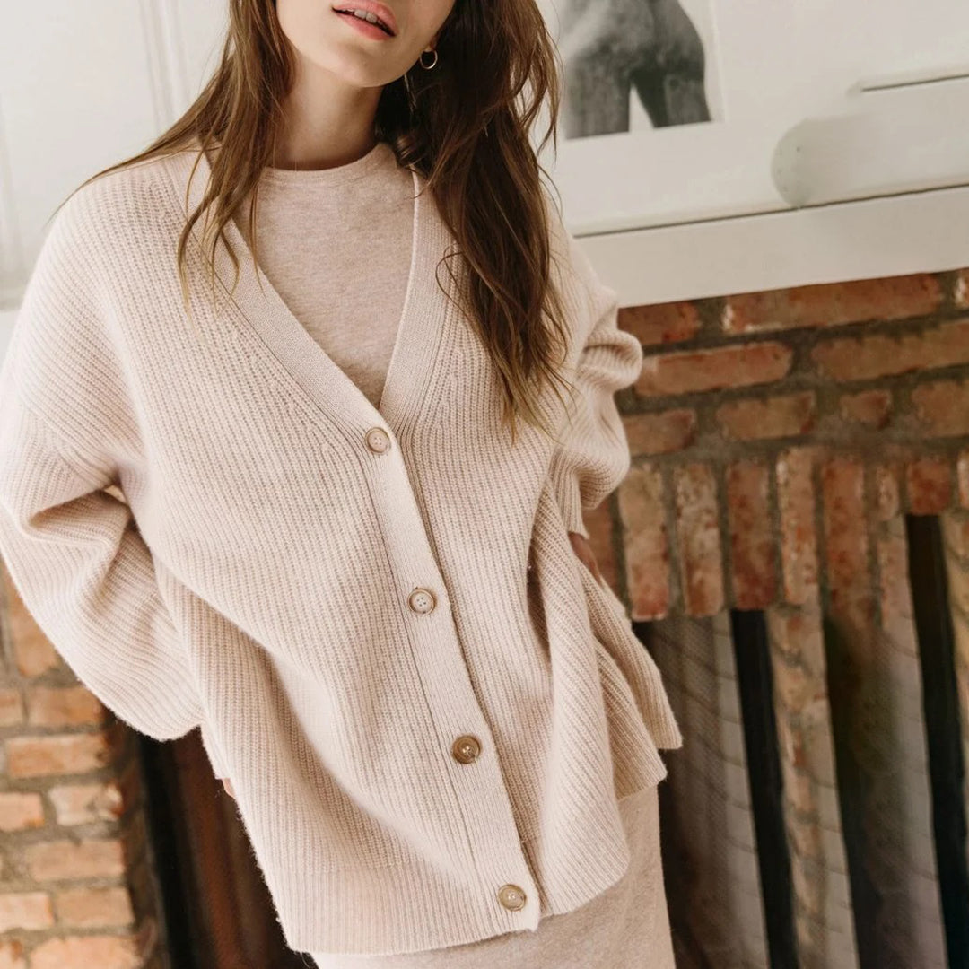 Donna con cardigan beige in maglia, davanti a un camino in mattoni. Moda autunnale, abbigliamento casual elegante, stile accogliente.