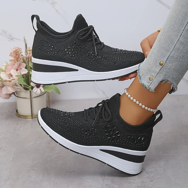 Scarpe da ginnastica nere con zeppa, design traforato, indossate con jeans chiari. Sfondo con fiori decorativi. Moda casual donna.