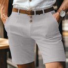 Uomo in pantaloncini grigi eleganti, camicia bianca, cintura marrone, orologio al polso. Moda estiva maschile, abbigliamento casual chic.