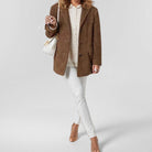 Donna in giacca di pelle marrone, maglione bianco, pantaloni bianchi e sandali beige, con borsa a tracolla bianca. Moda autunnale elegante.