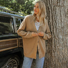 Donna con cardigan beige appoggiata a un albero accanto a un'auto d'epoca. Moda autunnale, stile casual, natura.
