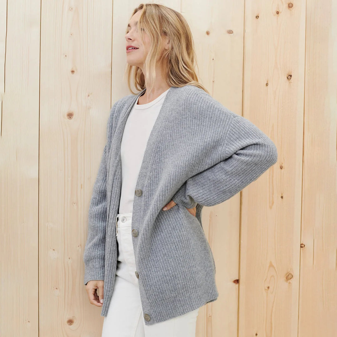 Donna con cardigan grigio in maglia, abbinato a maglietta bianca e jeans, su sfondo di pannelli in legno chiaro. Moda casual autunnale.
