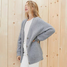 Donna con cardigan grigio in maglia, abbinato a maglietta bianca e jeans, su sfondo di pannelli in legno chiaro. Moda casual autunnale.