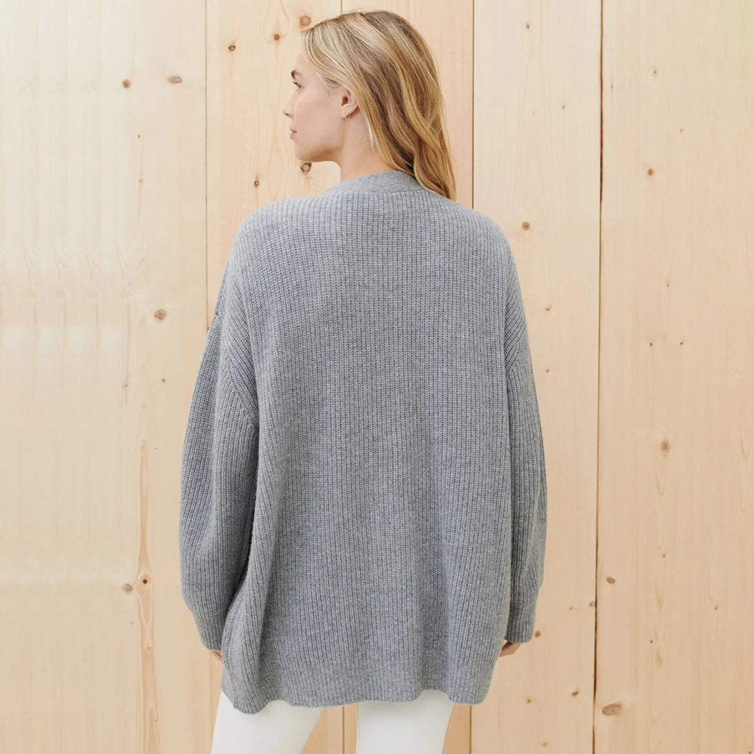 Donna di spalle con maglione grigio oversize in maglia a coste, sfondo in legno chiaro. Moda autunnale, abbigliamento casual elegante.