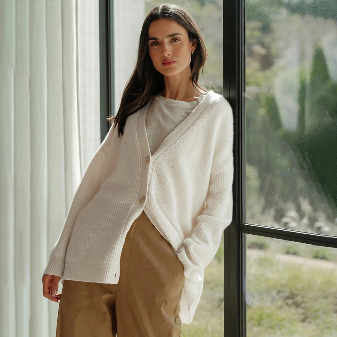 Donna con cardigan bianco in maglia, pantaloni beige, davanti a una finestra luminosa. Moda autunnale, abbigliamento casual elegante.