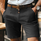 Uomo in pantaloncini eleganti neri con cintura marrone e camicia bianca, orologio al polso, moda estiva maschile, abbigliamento casual chic.