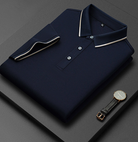 Camicia polo blu scuro piegata con colletto a contrasto, abbinata a un elegante orologio da polso nero su sfondo grigio. Moda uomo, abbigliamento casual.